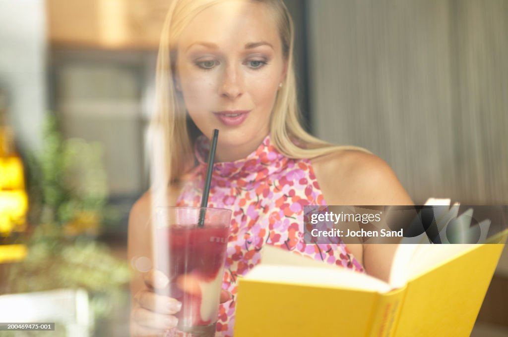 Young woman reading book in bar, view through window