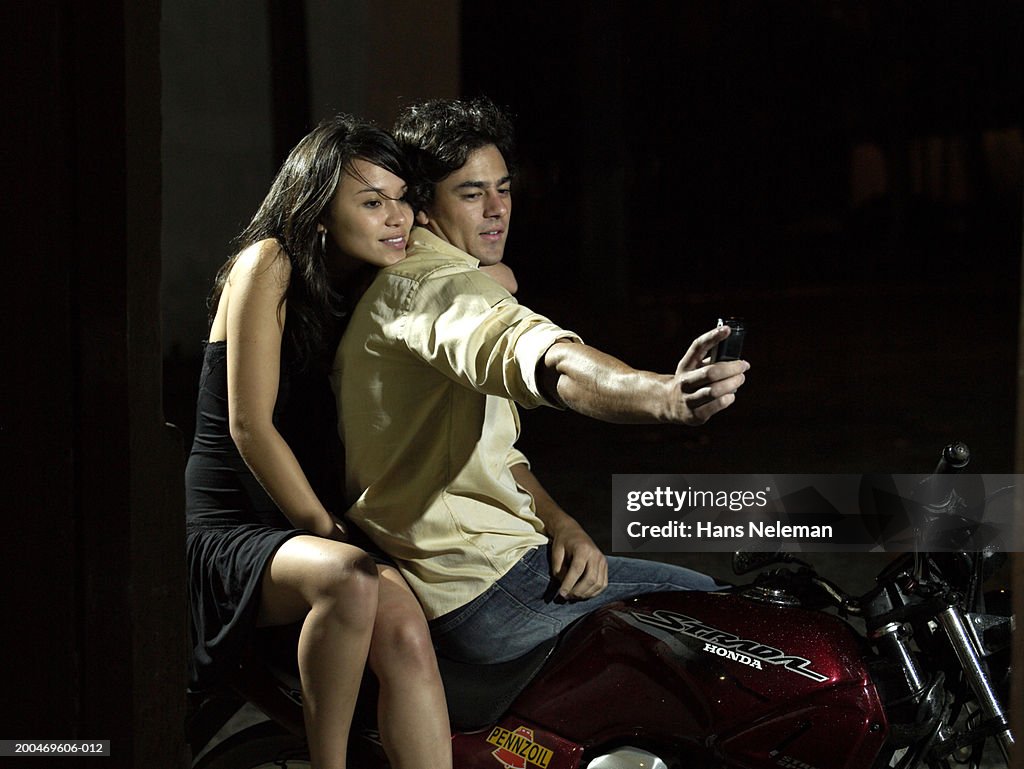 Young couple on motorcycle, man taking photo with mobile phone, night