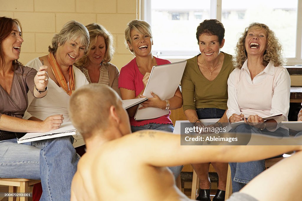 Mature women in life drawing class, smiling and laughing at male model