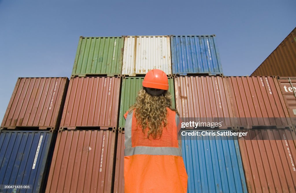 Woman wearing hard hat standing by containers in dock, rear view