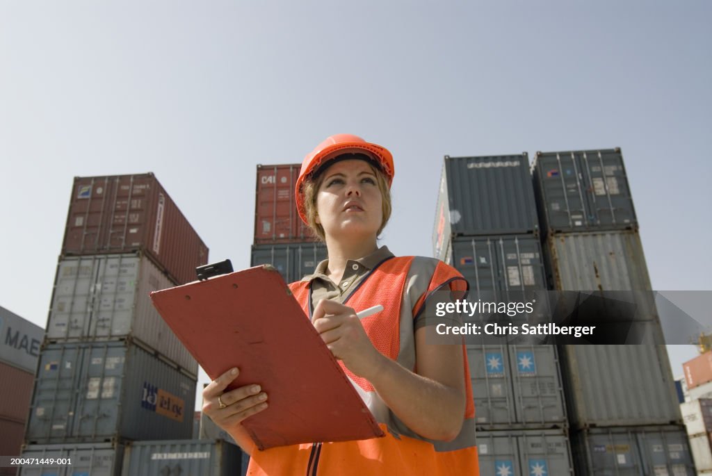 Woman wearing hard hat standing by shipping containers with clipboard