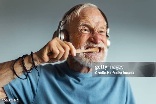 senior man brushing teeth in bathroom, listening music with wireless headphones. portrait of older man with healthy, gray hair. - zahnkaries stock-fotos und bilder