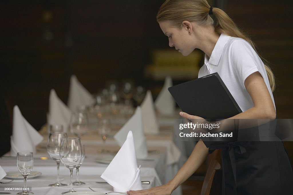 Young waitress adjusting table settings in restaurant, side view