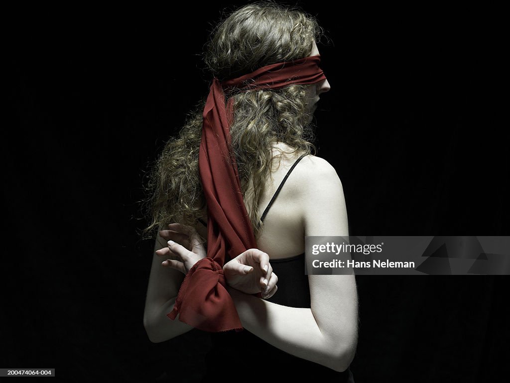 Young woman wearing blindfold, arms tied behind back, rear view
