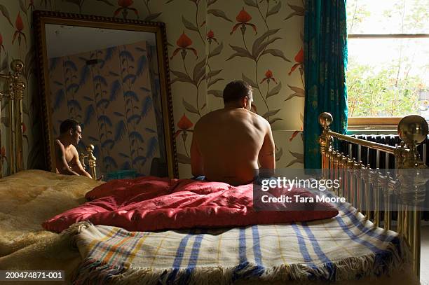 young man sitting at edge of bed, reflection in mirror, rear view - bedroom mirror stock-fotos und bilder