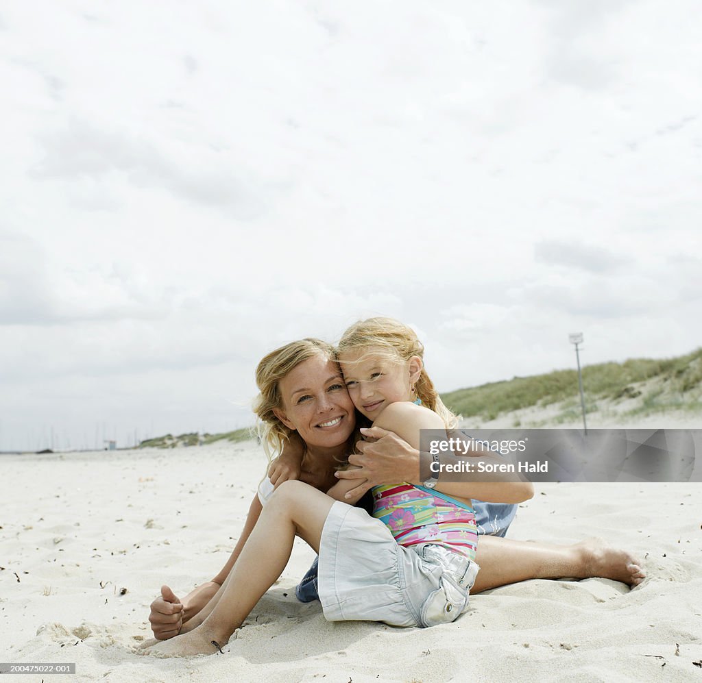 Mother embracing daughter (6-8) on sandy beach, smiling, portrait
