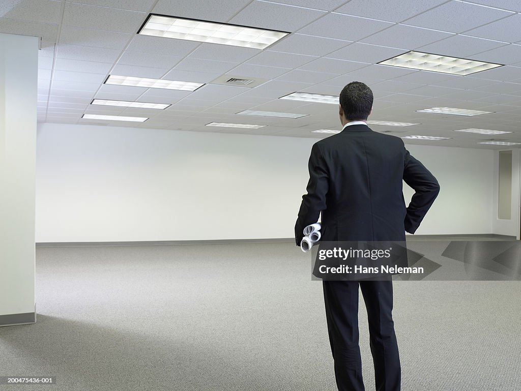 Businessman with rolls of plans, standing in empty office, rear view