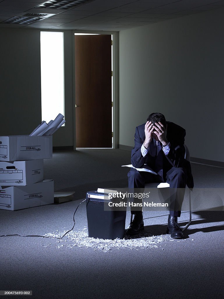 Businessman covering face, sitting by shredder in darkened room
