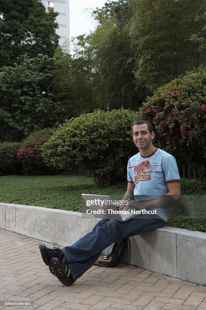 Young man with laptop sitting on concrete wall, smiling, portrait