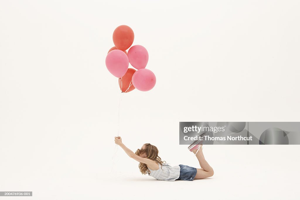 Girl (8-10) lying on stomach, holding bunch of balloons, side view