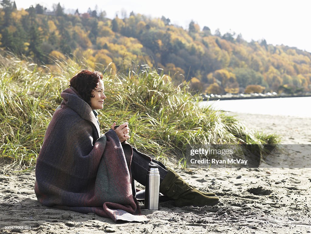 Mature woman sitting on beach, wrapped in blanket, autumn, side view