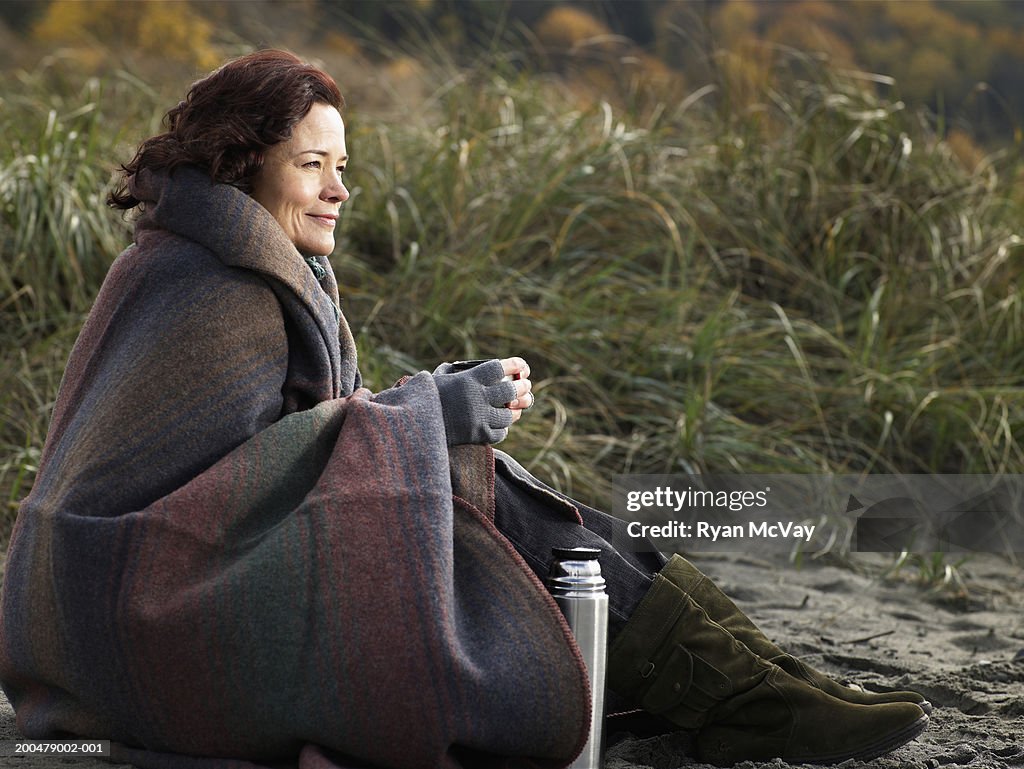 Mature woman sitting on beach, wrapped in blanket, autumn, side view