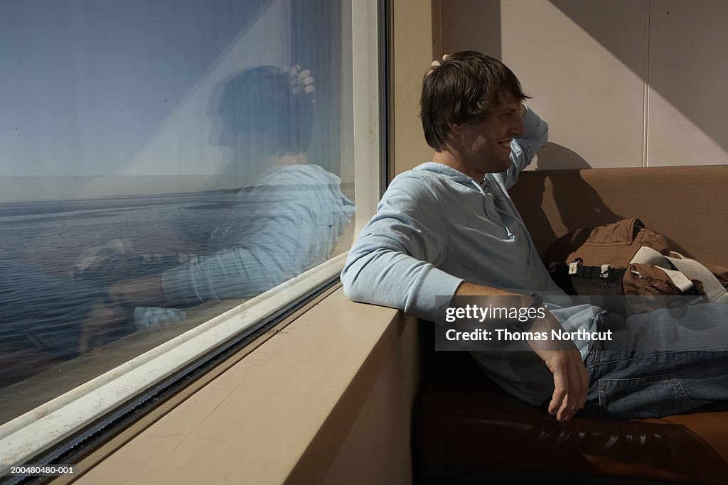 Young man leaning against window sill on ferry, laughing, side view