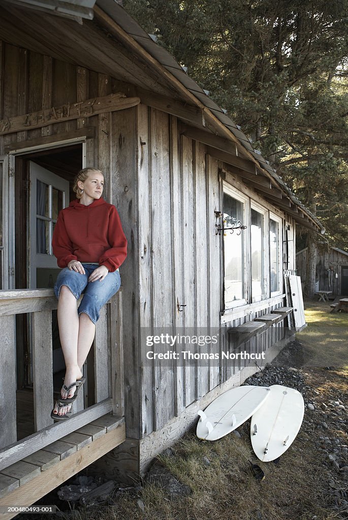 Young woman sitting atop railing on porch of cabin, looking away