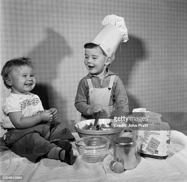 Boy wearing a chef's hat stirs ingredients in a bowl as a toddler looks on, November 1955.