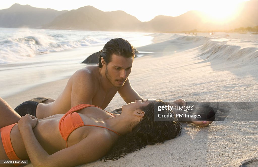 Young couple lying on beach, looking at each other, sunset