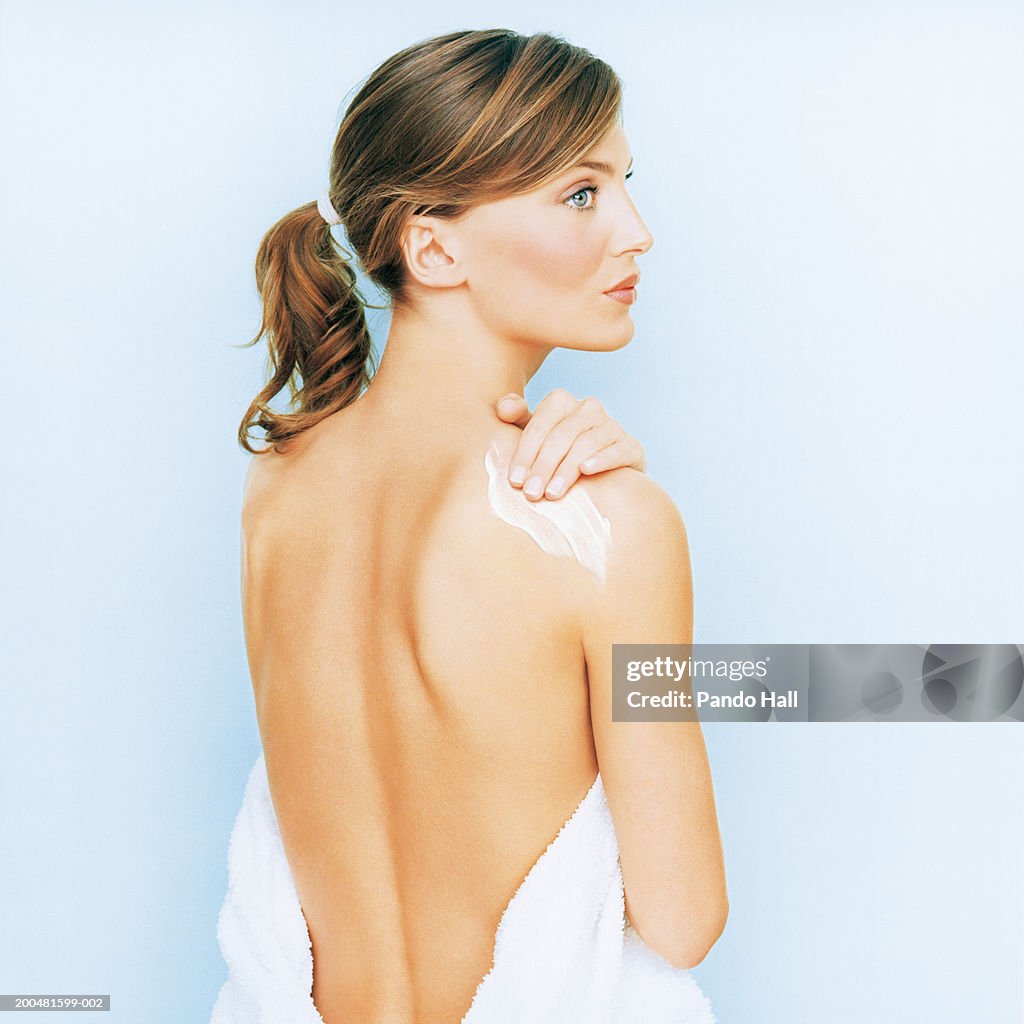Young woman rubbing cream on shoulder, wearing towel, side view
