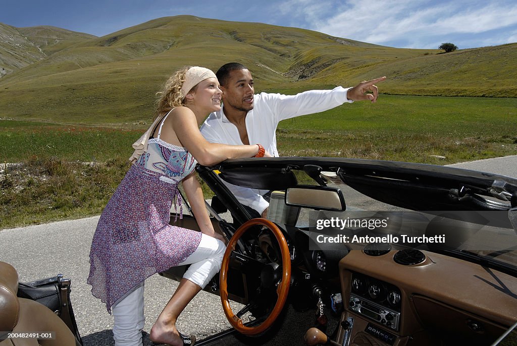 Young man and woman beside convertible car, man pointing