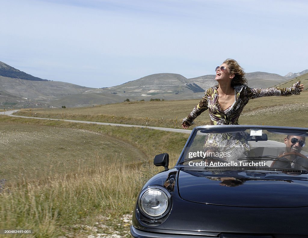 Man and woman riding in convertible car, woman has arms outstretched