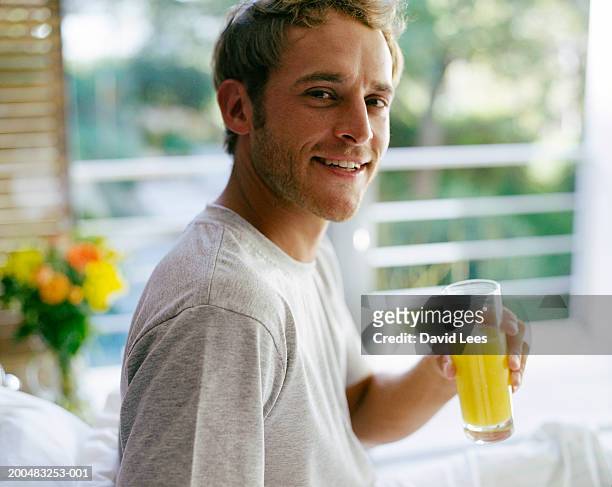 young man sitting on bed, holding glass of orange juice, portrait - orange juice stock-fotos und bilder