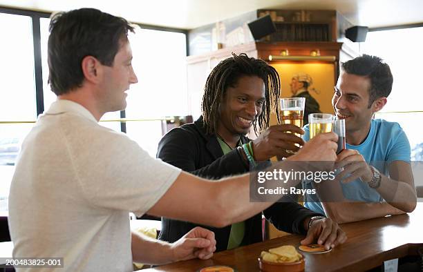 three men drinking at bar, smiling and raising glasses - bar man t shirt stock-fotos und bilder