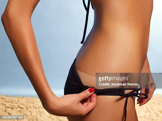 young woman pulling down bikini bottom to reveal tan line, close-up - braun stock-fotos und bilder