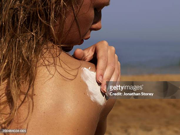 young woman applying sun lotion to shoulder, on beach, rear view - applying stockfoto's en -beelden