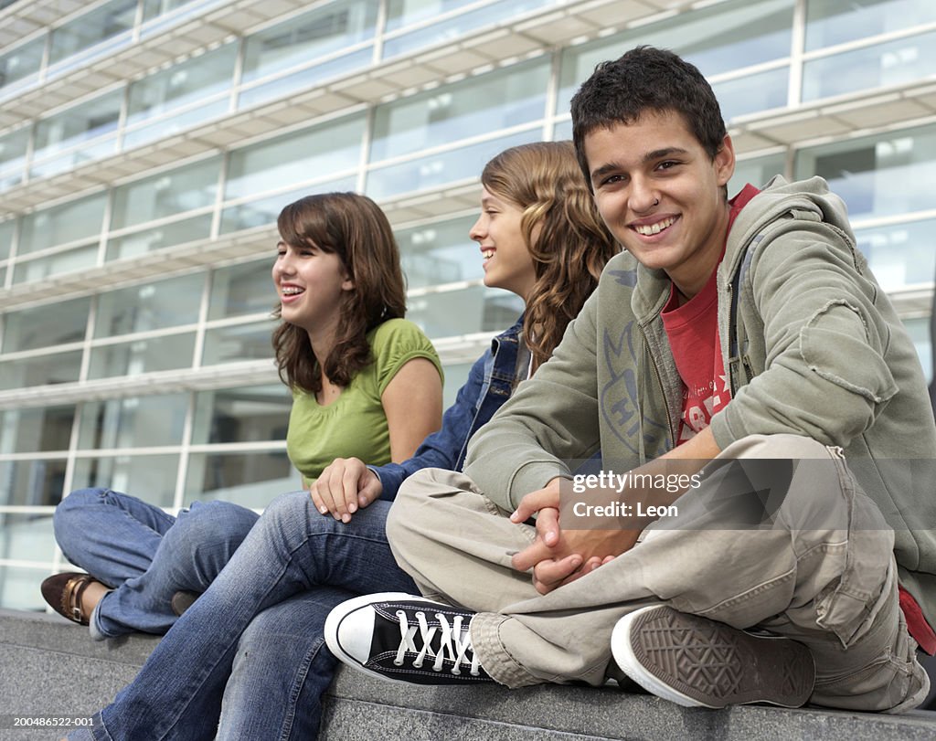 Teenage boy (16-18) sitting on wall with two teenage girls, smiling