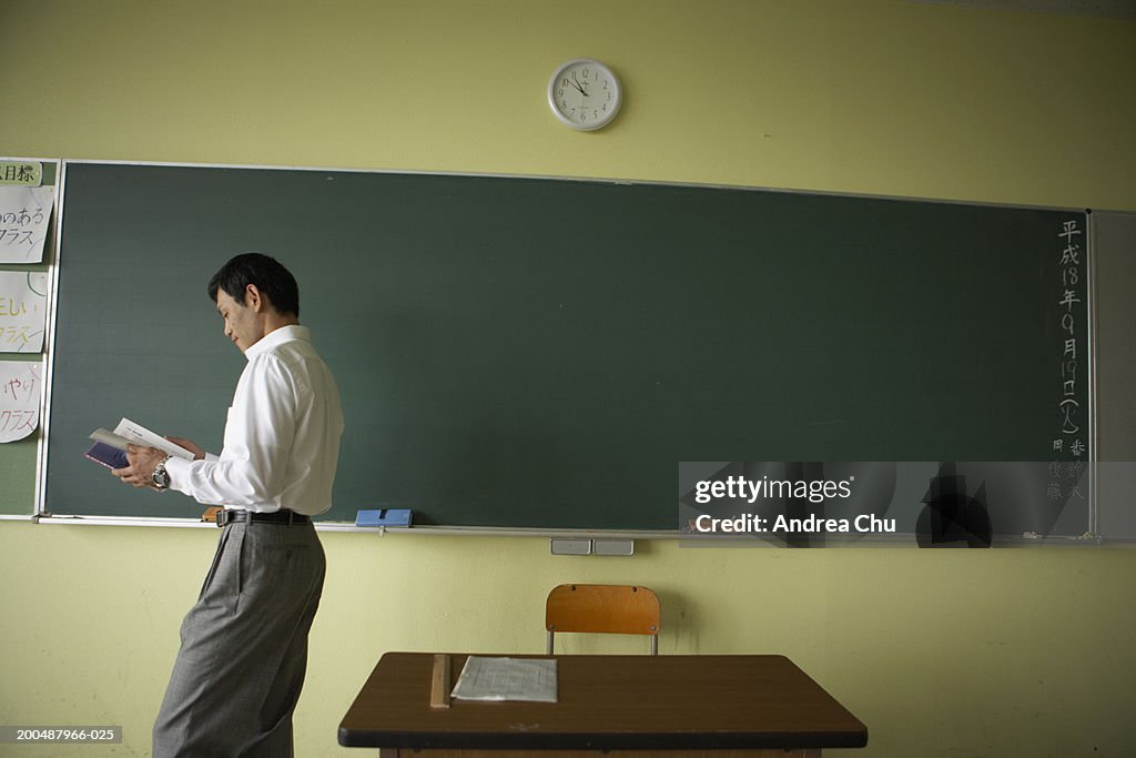 Teacher in front of blackboard, reading book, side view