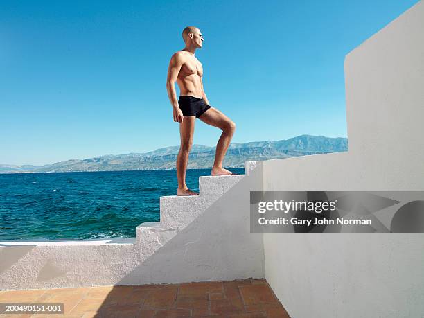 young man standing on white steps by sea - men swimwear stock pictures, royalty-free photos & images