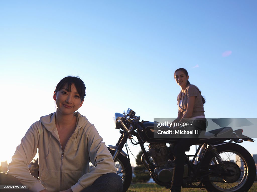 Two young women, one sitting on lawn, one sitting on motorcycle