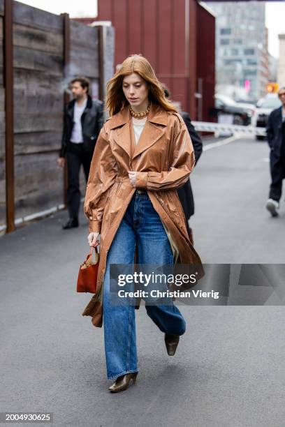 Guest wears brown vinyl coat, denim jeans, red bag outside Ulla Johnson on February 11, 2024 in New York City.