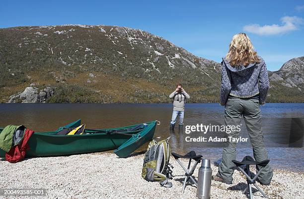 man in lake taking photograph of woman on shore - knöcheltief im wasser stock-fotos und bilder
