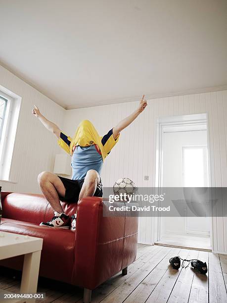 young man sitting on back of sofa, t-shirt pulled over head, cheering - danish sports stock pictures, royalty-free photos & images