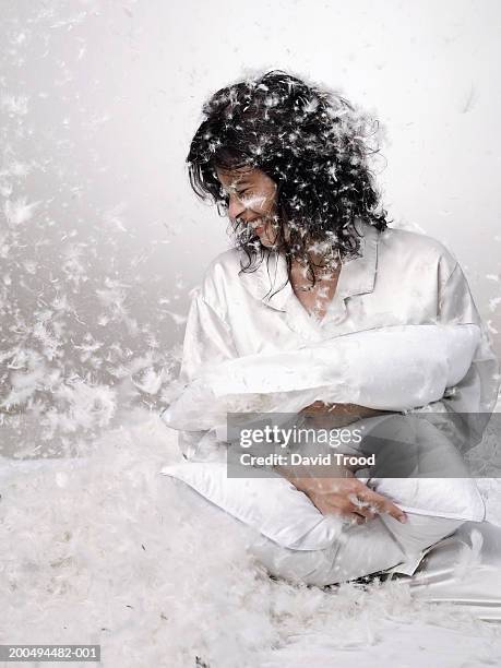 young woman clutching pillow, surrounded by falling feathers, laughing - falling feathers stock pictures, royalty-free photos & images