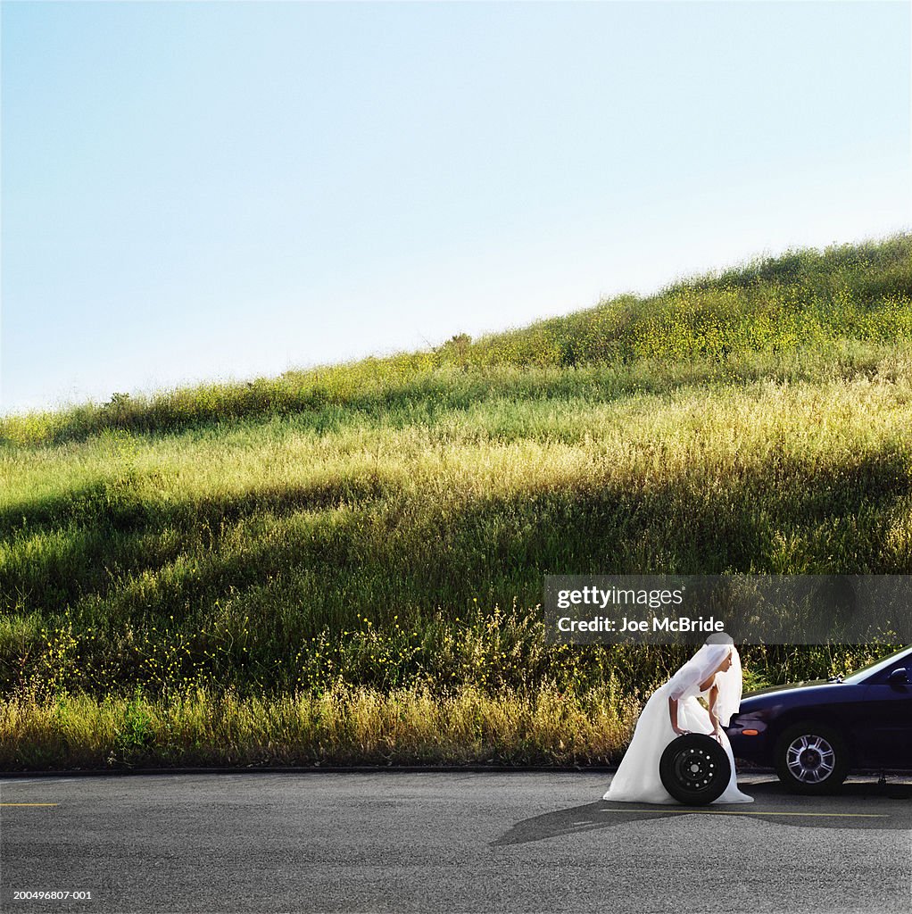Bride rolling car wheel on side of road, side view