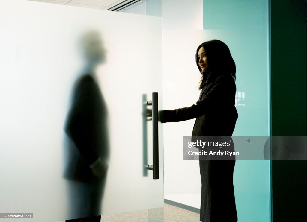 Businessman standing behind frosted glass, woman entering office