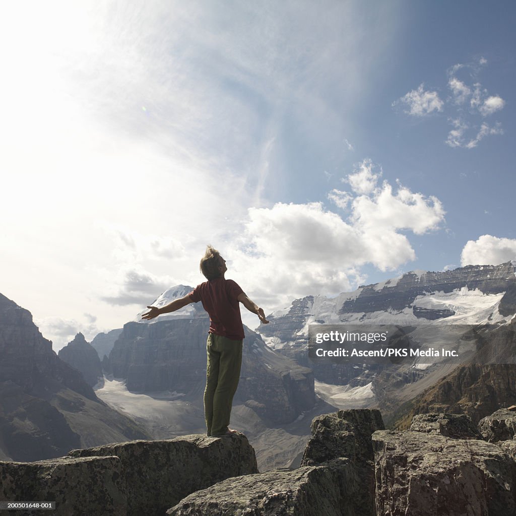 Mature male hiker atop rock in mountains, arms outstretched, rear view