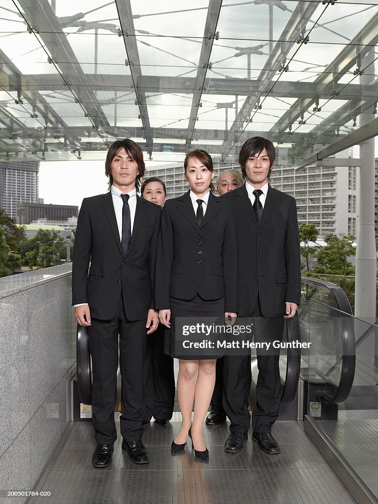 Five executives standing at top of escalator, portrait