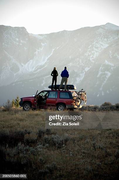 young couple standing on suv roof, looking at mountain view, rear view - suv stock-fotos und bilder