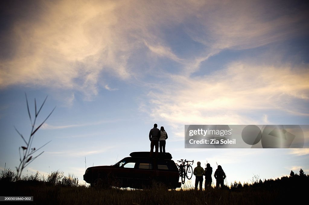 Young group watching sunset beside SUV, couple on car roof, rear view