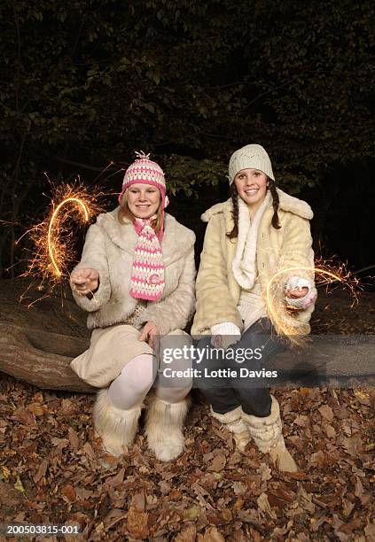 two teenage girls (13-14) sitting on log, playing with sparklers - autumn friends coats stock pictures, royalty-free photos & images