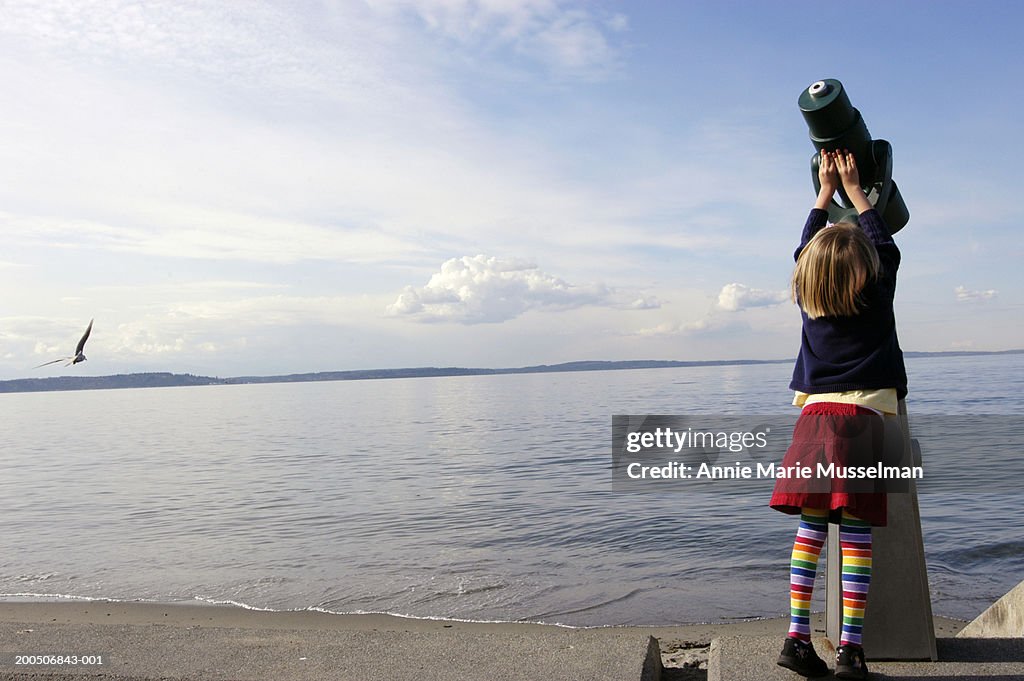 Girl (5-7) next to coin-operated binoculars beside sea, rear view