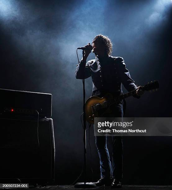 young man with electric guitar singing into microphone on dark stage - zangeres stockfoto's en -beelden