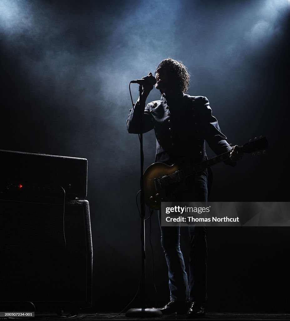 Young man with electric guitar singing into microphone on dark stage