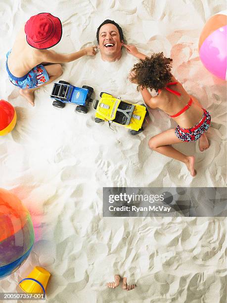 boy and girl (5-7) beside father buried in sand, overhead view - buried in sand stock-fotos und bilder