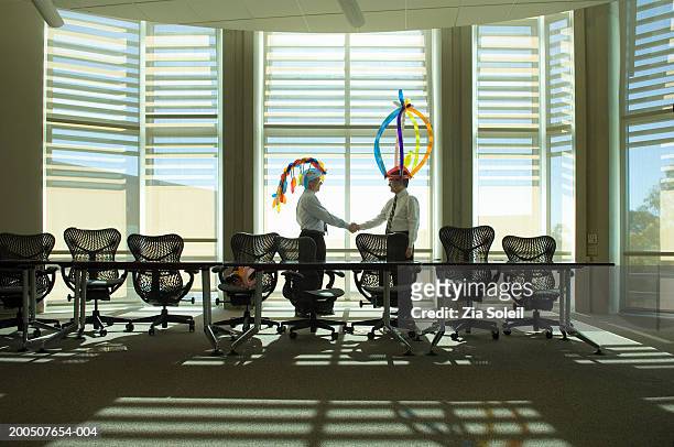 two mature businessmen wearing balloon hats, shaking hands in office - soleil humour stockfoto's en -beelden
