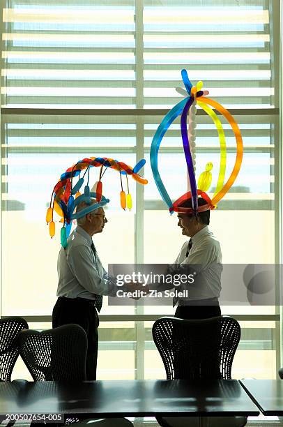 two mature businessmen wearing balloon hats, shaking hands in office - soleil humour stockfoto's en -beelden