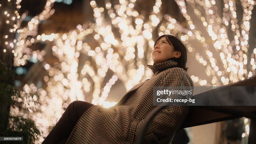Portrait of Asian woman sitting under Christmas illumination lights in city at night
