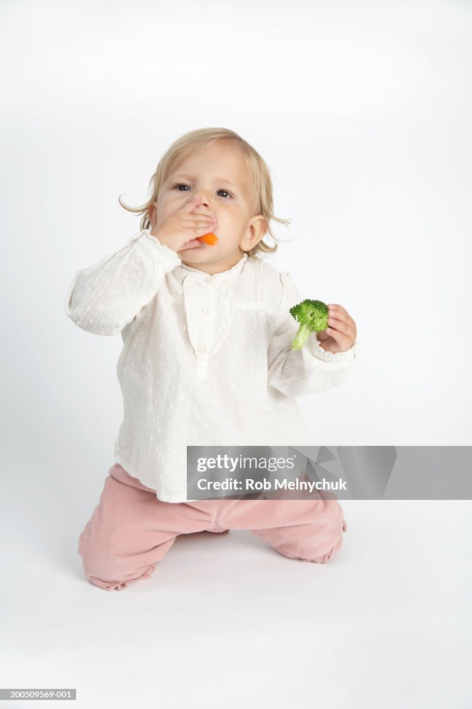 Baby girl (15-18 months) eating carrots and broccoli, looking up